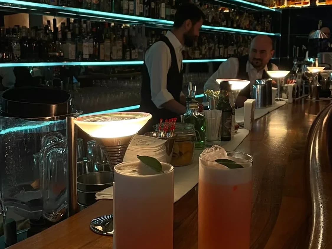 Bartresen mit zwei rosa Longdrinks, beleuchtetem Flaschenregal und Barkeepern im Hintergrund